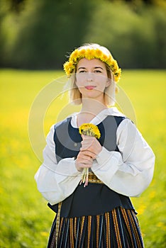 Young woman in national clothes wearing yellow dandelion wreath in spring field. Ligo