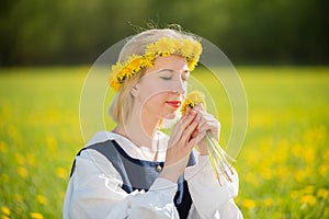 Young woman in national clothes wearing yellow dandelion wreath in spring field. Ligo