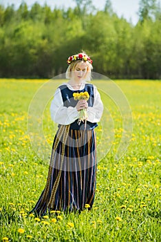 Young woman in national clothes wearing yellow dandelion wreath in spring field. Ligo