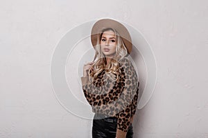 Young woman with natural make-up in stylish leopard sweater in fashionable black leather pants in stylish hat posing