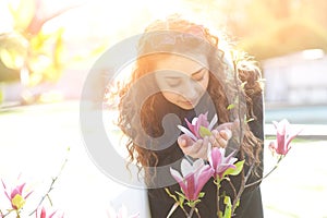 Young woman near blossoming magnolia flowers tree in spring park on sunny day. Beautiful happy girl enjoying smell in a flowering