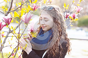 Young woman near blossoming magnolia flowers tree in spring park on sunny day. Beautiful happy girl enjoying smell in a flowering