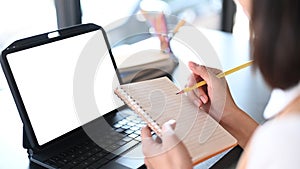 Young woman office worker holding pen writing on empty notebook in front of computer tablet.