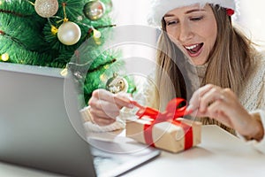 Young woman opening Christmas or new year present when calling to friends via video chat from home