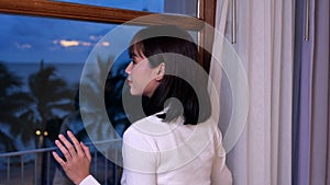 Young woman opening curtain and standing in apartment home. Thai female looking through window to see sea view on sunset.