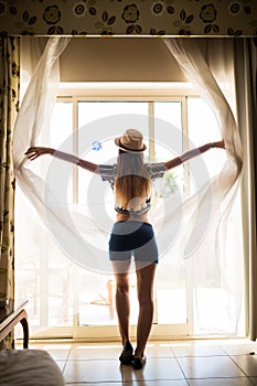 Young woman opening curtains in a bedroom at hotel room before to go outdoors