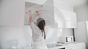 Young woman opens the door and takes out glass cups from cabinet. She in white T-shirt and jeans stretches arms to the