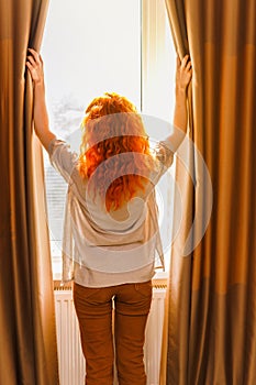 Young woman with orange hair stand in the room opening curtain seeing the beautiful sunrise.