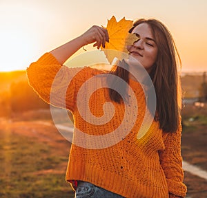 Young woman in a orange sweater with with yellow leaves, outdoor portrait in soft sunny daylight. Autumn. Sunset. Cozy