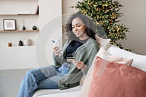 Young woman ordering gift during Christmas holiday at home using smartphone and credit card. Shopping online during holidays