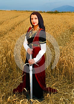 Young woman with ornamental dress and sword in hand standing on a wheat field with sunset. Natural background..