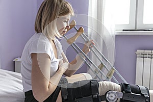 A young woman with an orthosis on her leg is sitting on the bed. Tries to get up with crutches. Postoperative period