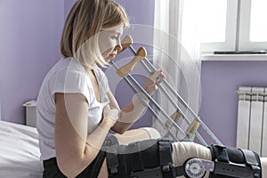 A young woman with an orthosis on her leg is sitting on the bed. Tries to get up with crutches. Postoperative period photo