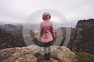 Young woman in outdoor clothing standing on cliff`s edge enjoying view of mountain ridge and valley with wild forest