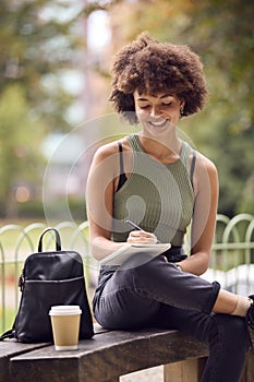 Young Woman Outdoors In Park Sitting On Bench Working Writing In Notebook 