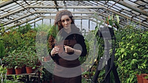 Young woman owner of hothouse is recording video about gardening for her videoblog standing in greenhouse and holding