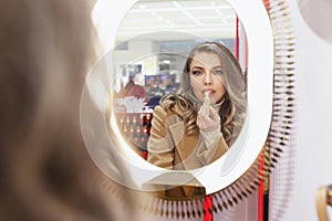 Young woman paints lips with lipstick in front of a mirror in a cosmetics store. Beautiful blonde in a beige coat. Fashion, style