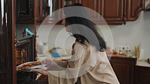 A young woman in pajamas opens the oven and takes out a slightly heated pizza in an aluminum form for baking with her