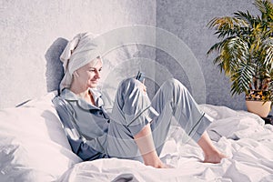 Young woman in pajamas and a towel on her head sits in bed and looks at her phone