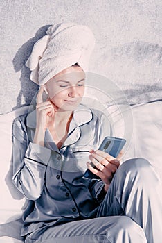 Young woman in pajamas and a towel on her head sits in bed and looks at her phone