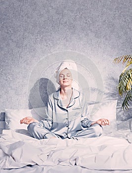 A young woman in pajamas and a towel on her head sits in the lotus position on the bed the morning sun shines on her