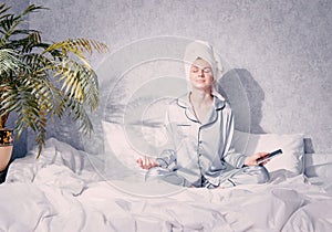 A young woman in pajamas and a towel on her head sits in the lotus position on the bed the morning sun shines on her