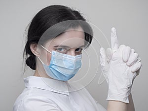 Young woman patient in a medical mask puts on protective surgical sterile gloves on her arm, on gray background, protection