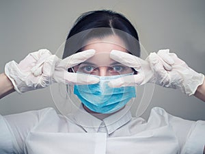 Young woman patient in a medical mask puts on protective surgical sterile gloves on her arm, on gray background, protection