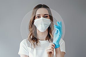 Young woman patient in a medical mask puts on protective surgical sterile gloves on her arm, isolated on gray background,