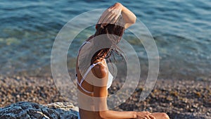 Young woman with perfect tanned slim body in white bikini relaxing on stone at pebble beach