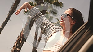 Young woman performs dance movements leaning out of car window traveling along road with palm trees