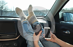 Young woman with phone in warm socks holding her legs on car dashboard and drinking coffee. Cozy atmosphere