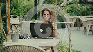 A young woman photographer drinks a cocktail while working with her laptop in an outdoor cafe
