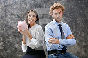 Young woman with piggy bank and her displeased husband on grey background