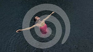 Young woman in pink bikini floating on the back in the dark water of simming pool, ocean or lake under the rain drops