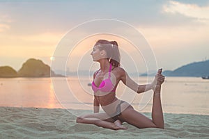 Young woman in pink bra and black panties sitting on beach stretching her legs during sunset at sea. Fitness girl doing