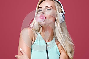 Beautiful blonde woman blowing pink bubble gum. Girl listen music in headphones. Fashion and lifestyle portrait.