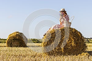 A young woman in pink dress and a straw hat is sitting on a haystack.