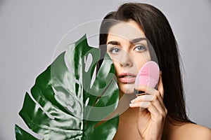 Young woman with pink face exfoliator brush silicone cleansing device for sensitive normal skin with tropical palm leaf on gray