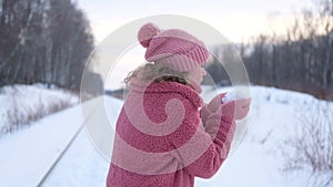 Young woman in pink faux fur coat blows snow from palms in winter