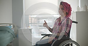 Young woman with pink hair sit in wheelchair and shows thumbs up.