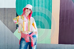 Young woman with pink hair and sunglasses in Bucket hat and multicolor strippled shirt posing on graffiti wall