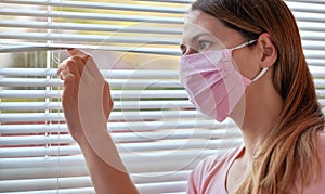 Young woman in pink home made cotton virus face mask, looking through window blinds outside. Quarantine or stay at home during