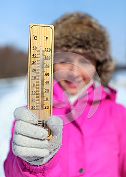 Young woman in pink jacket holds wooden thermometer outside on sunny but cold day, illustrating weather with freezing temperatures