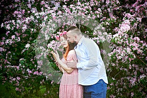 Young woman in a pink lace dress and a bearded adult man in a white shirt hold hands against the background of apple trees in