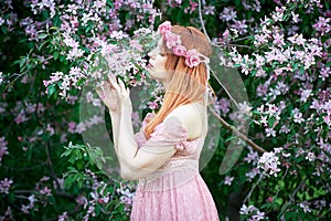Young woman in a pink lace dress and a wreath of roses inhales the fragrance of a flowering tree. An idea for a photo shoot