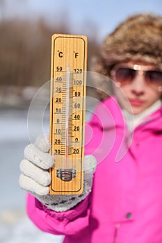 Young woman in pink winter jacked, gloves and furry hat holding thermometer that is showing -5 degrees. Winter / cold days coming