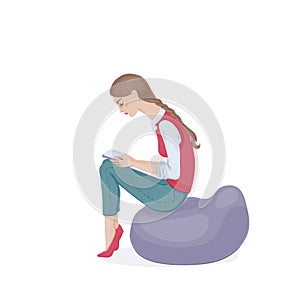 A young woman with a plait in a pink waistcoat and jeans sitting on a beanbag chair and reading a book. A student girl