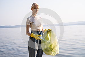 A young woman with a plastic garbage bag stands by the lake and looks into the camera. A call to clean up the