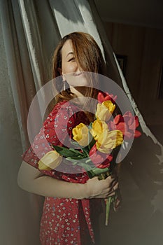 Young woman is playful natural beauty in red dress with floral print holding lush fresh blooming red and yellow tulips in light